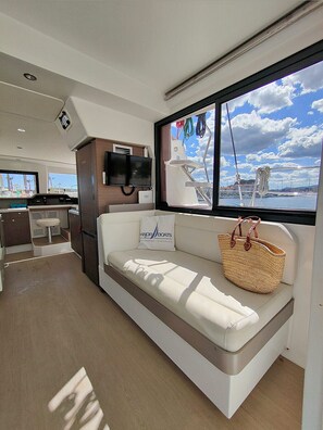 A boat interior with a sofa, a television, a window with a view of the sea, and a small kitchen area.