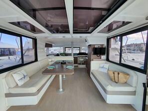 A boat interior with a dining area, seating, a kitchen, and large windows.