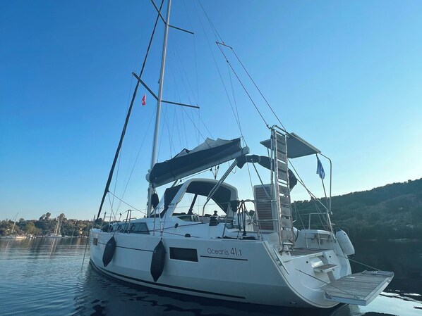Een zeilboot genaamd 'Oceanis 41.1' afgemeerd in een jachthaven met rustig water, een heldere lucht en een heuvelachtig landschap op de achtergrond.
