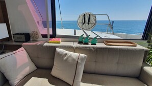 Een knus vakantiehuisje met een zachte zithoek, een kleine tafel met een lamp, een radio en decoratieve objecten, met uitzicht op een heldere blauwe zee.