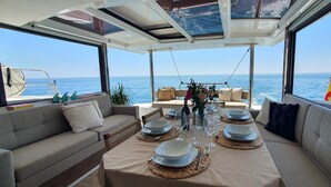 Een eethoek op een boot met een gedekte tafel voor twee, omringd door comfortabele zittingen en grote ramen met uitzicht op de zee.