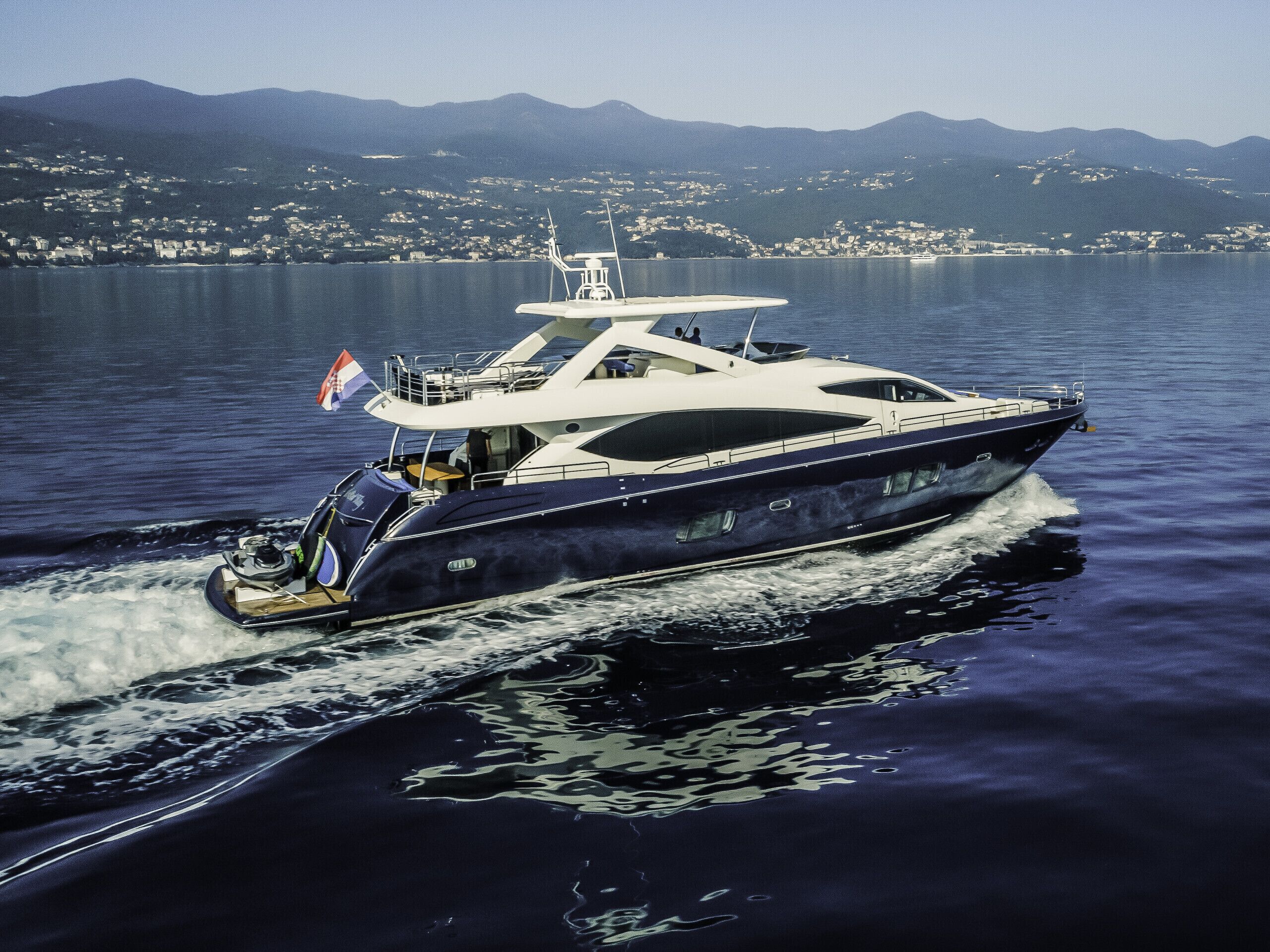 A large yacht on water with a flag, surrounded by a mountainous landscape.