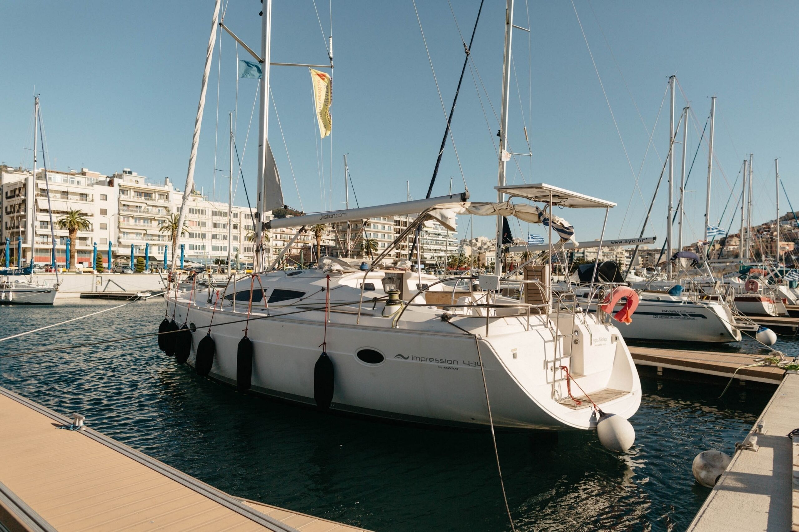 A marina with multiple yachts docked, a clear sky, and buildings in the background.