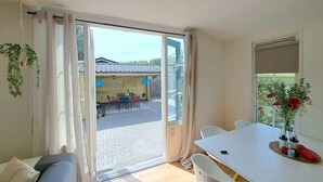 A room with a sliding glass door leading to an outdoor patio, a white dining table, a sofa, and a window with a plant. - Nature Base Near Fishing Waters (Heinkenszand)