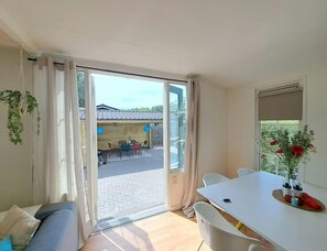 A room with a sliding glass door leading to an outdoor patio, a white dining table, a sofa, and a window with a plant. - Nature Base Near Fishing Waters (Heinkenszand)