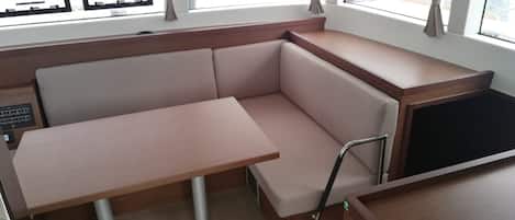 Uma cabine compacta com área de estar acolchoada, uma mesa pequena e vista para a marina através de grandes janelas.