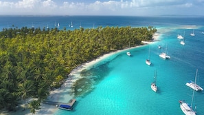 An aerial view of a tropical island with clear turquoise waters, a sandy beach, and numerous sailboats anchored near the shore.