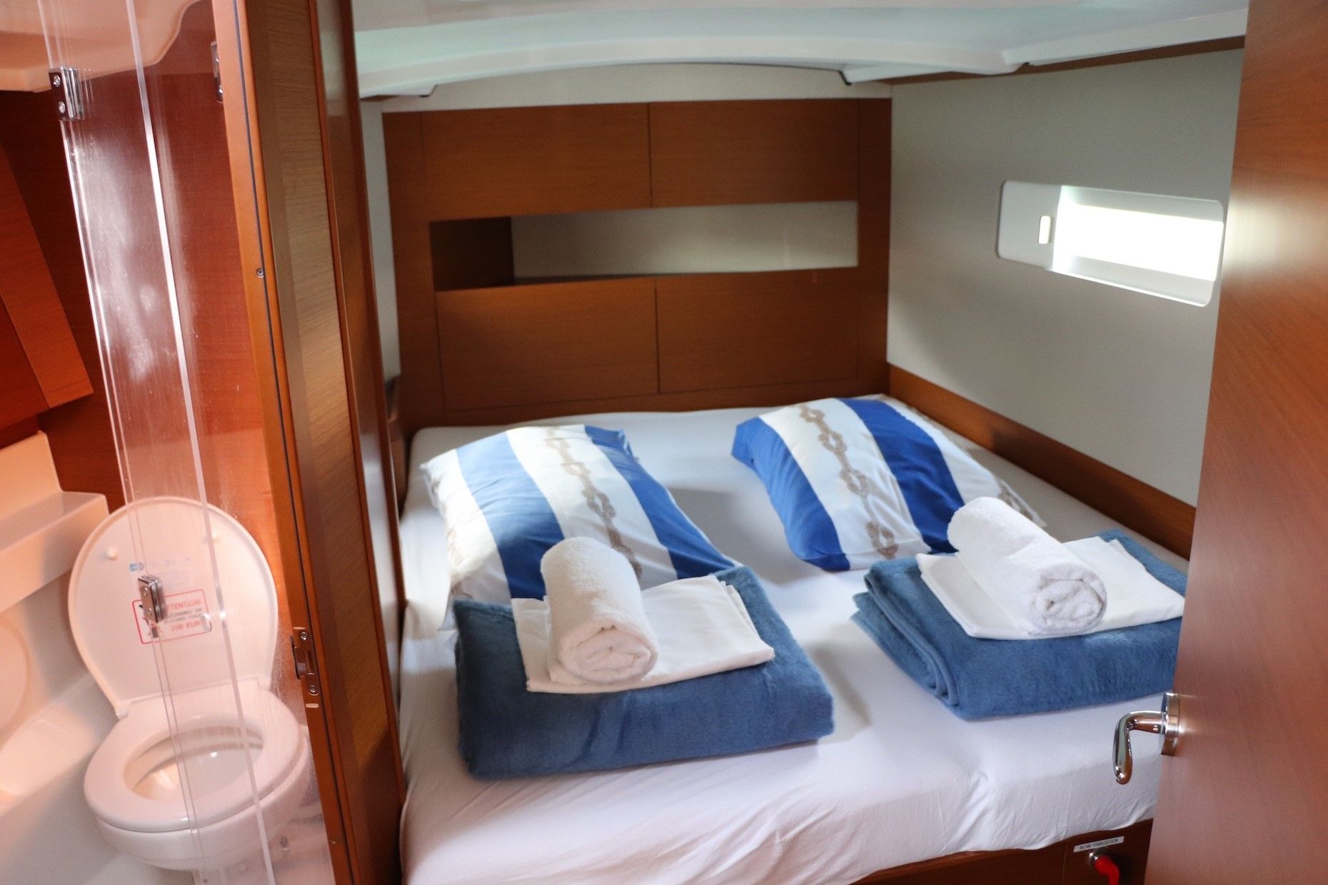A compact, well-lit cabin with a single bed featuring blue and white bedding, a wooden headboard, and a small window.