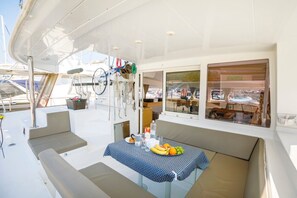 A yacht interior with a dining area, cushioned seating, a table with fruits and drinks, and a view of other yachts through the windows. - Sun-Kissed Sailing Adventure in Split, Croatia (Split)