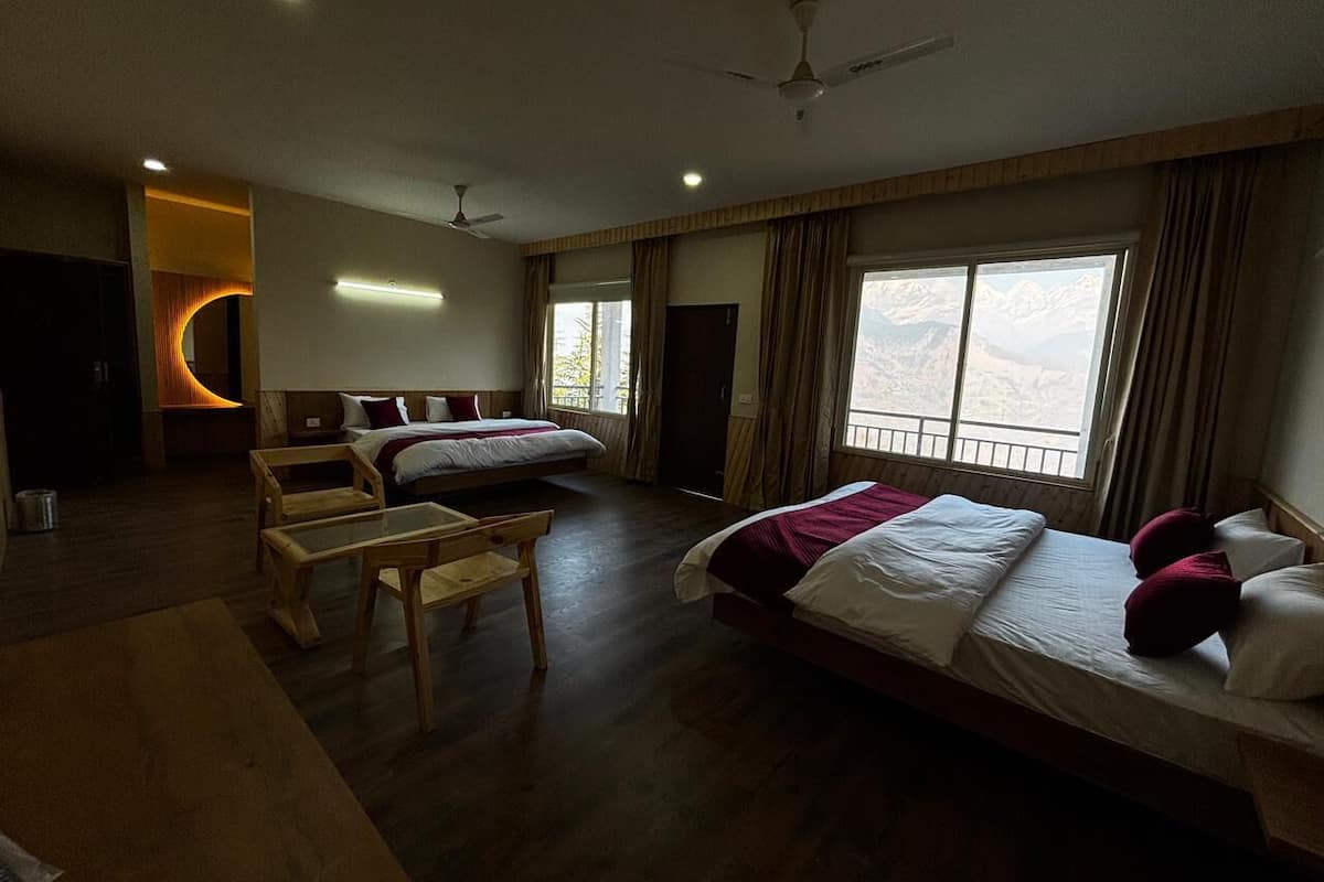 A hotel room with two beds, a wooden table, a ceiling fan, and large windows with a mountain view.