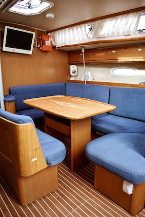 Un interior pequeño con una mesa de madera, asientos con cojines azules, un televisor montado y una ventana con cortinas.