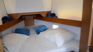 Una cama compacta con ropa de cama blanca y toallas azules, cabecera de madera y una pequeña estantería de madera.