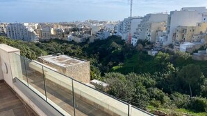 Terraço no terraço com guarda-corpo de vidro, vista de um horizonte urbano com edifícios e guindastes, e uma costa distante.