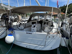 Eine Segelyacht namens 'BOBO' liegt in einer Marina, wo weitere Boote, ein Steuerrad und eine Winde sichtbar sind.