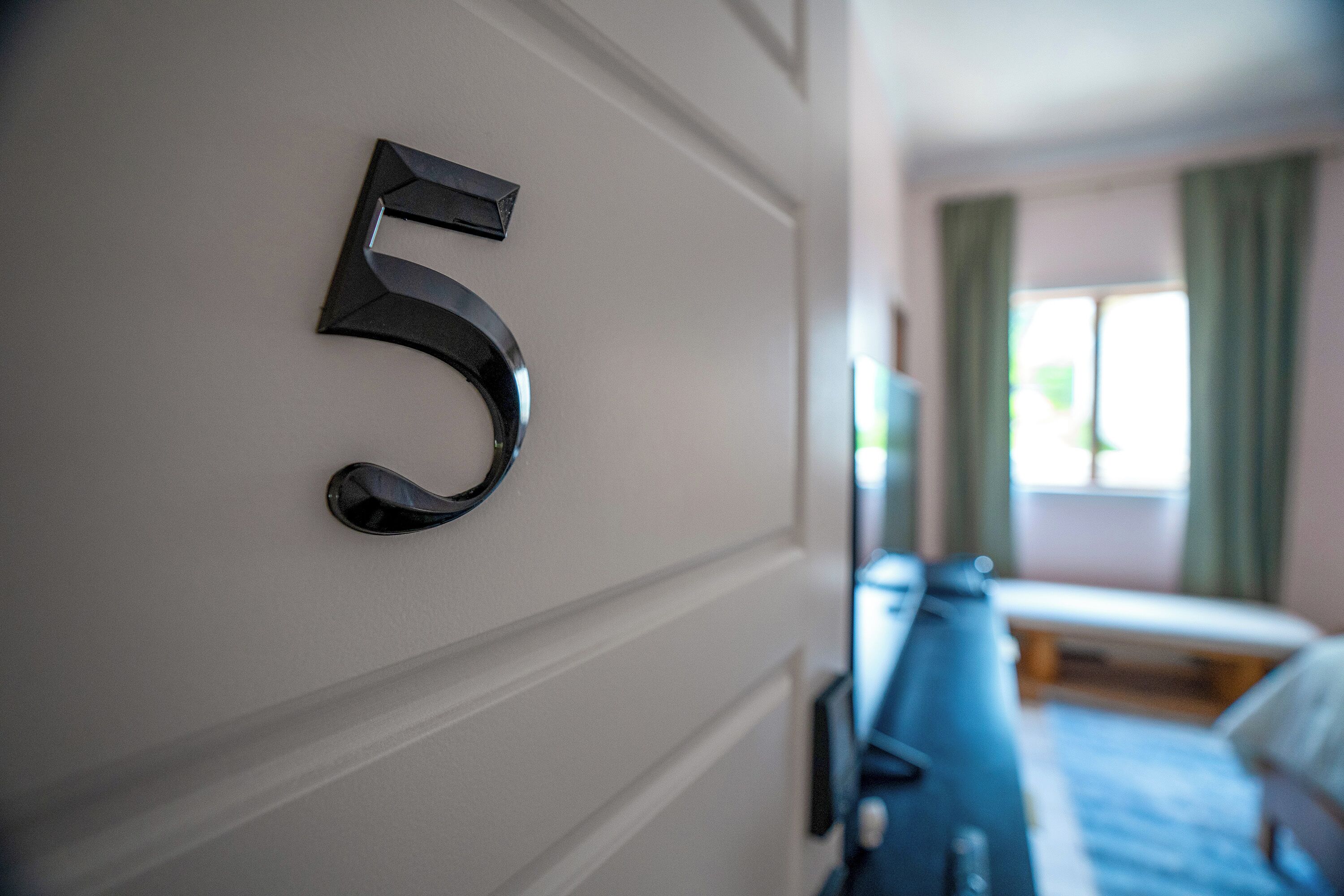 A hotel room with a number '5' on the wall, a bed, a TV, and a window with curtains.