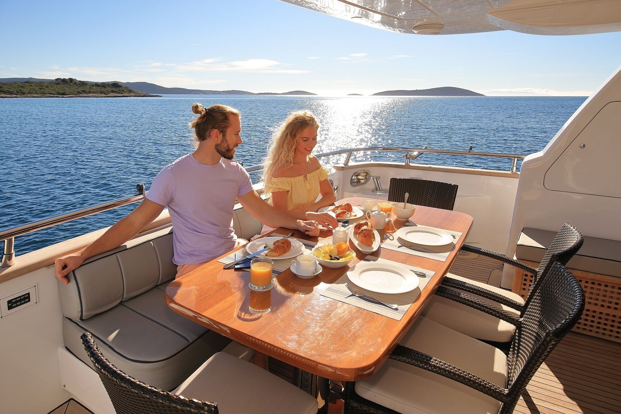 A couple dining on a boat with a table set for breakfast, surrounded by water and distant land.