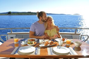 A couple dining on a boat with a table set for breakfast, including croissants, fruit, and juice.