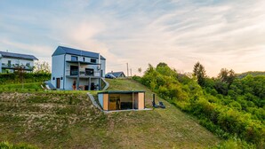 Ein modernes, zweistöckiges Haus mit Balkon, einem kleinen Anbau und einem grasbewachsenen Hügel.