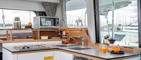 Een compacte keuken met een magnetron, spoelbak en kookplaat, met uitzicht op de jachthaven door grote ramen.