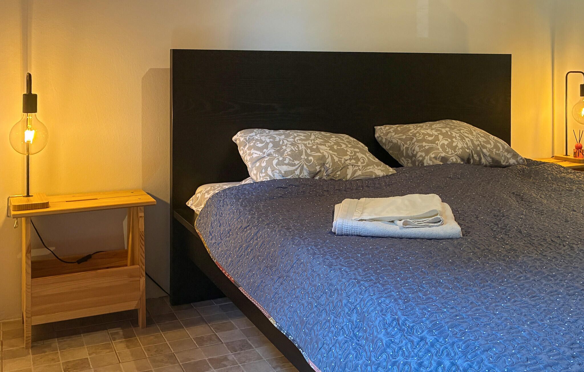 Una cama bien tendida con un edredón azul oscuro, dos almohadas estampadas y una toalla doblada. Se ve una mesita de noche con una lámpara, y un aplique en la pared.
