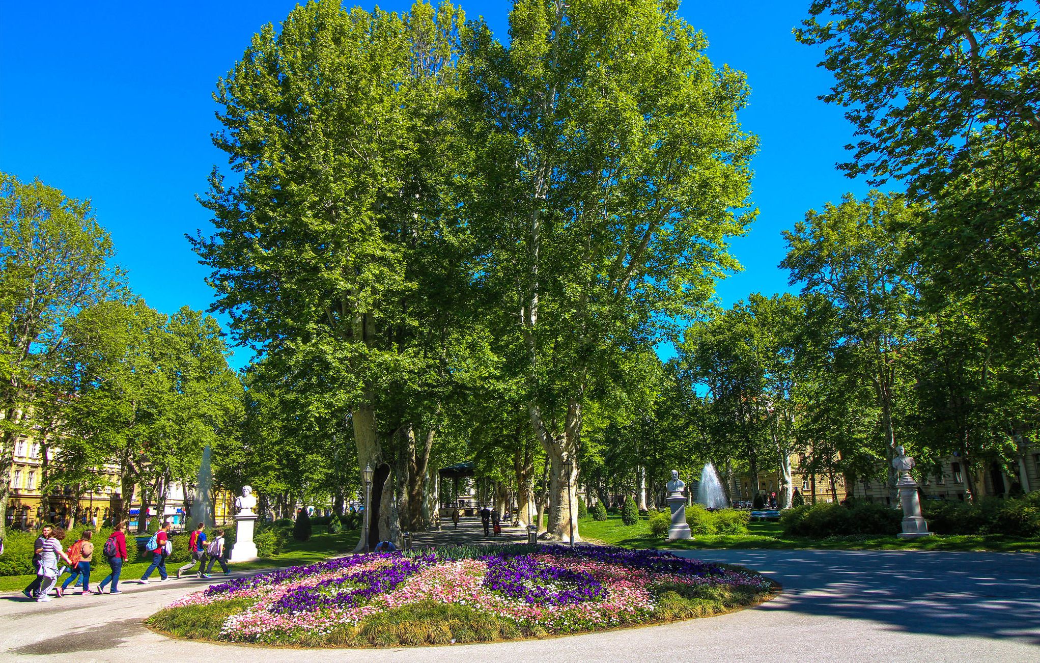 A park with a large tree, statues, and a flower bed.