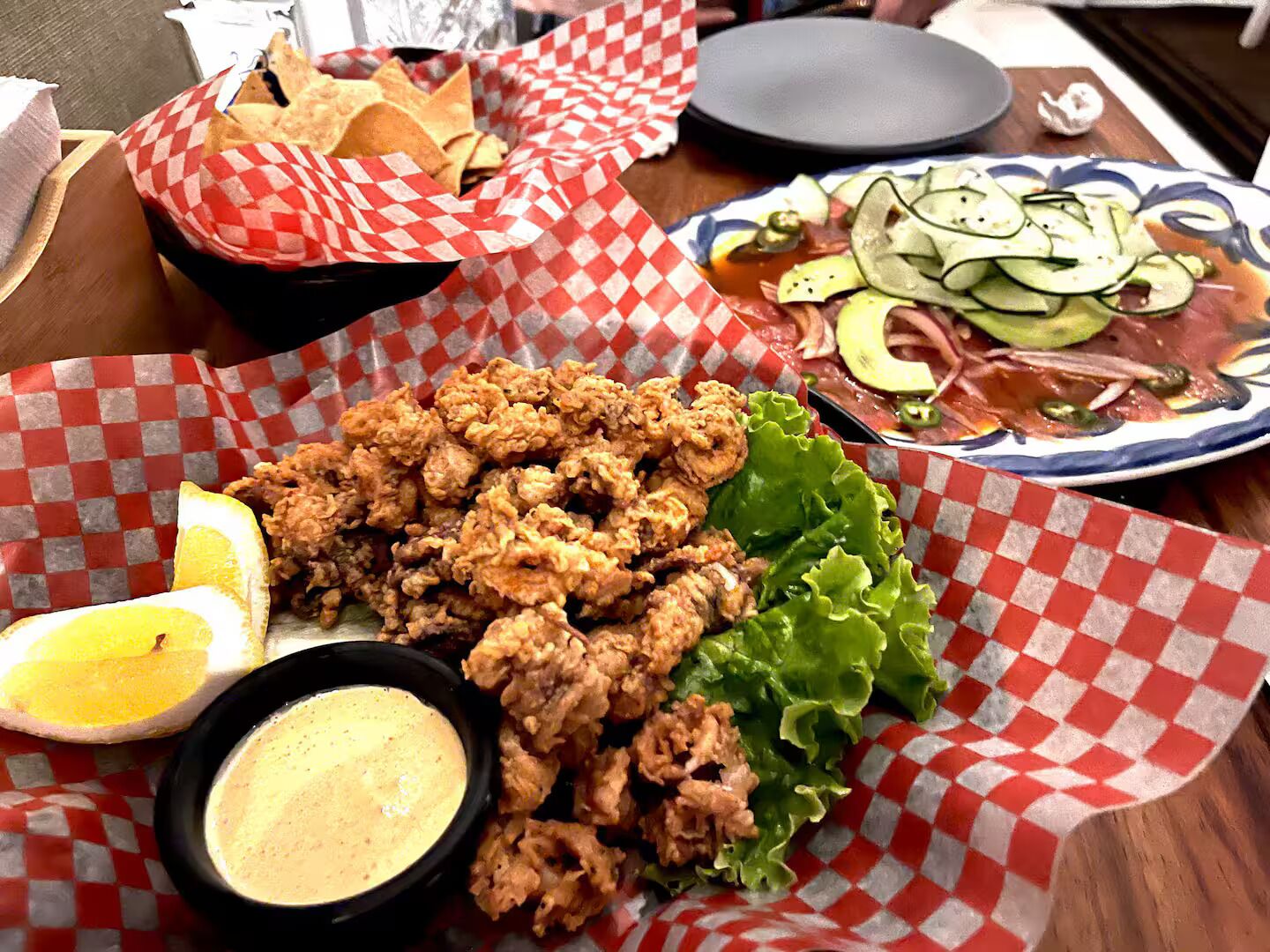A table with fried chicken, sliced cucumbers, lettuce, lemon wedges, and a dipping sauce.