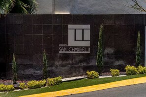 Een muur met een bordje voor hotel 'San Charbel II', omringd door groen en een gele stoeprand.