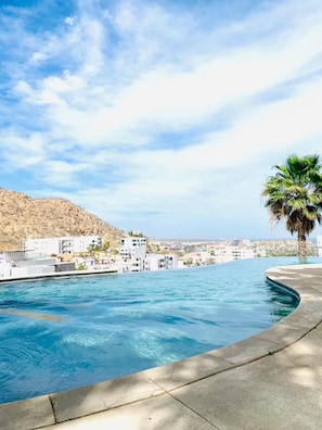 A swimming pool with a curved edge, a palm tree, and buildings in the background. - Getway with an incredible pool Marina Bay view! (Cabo San Lucas)