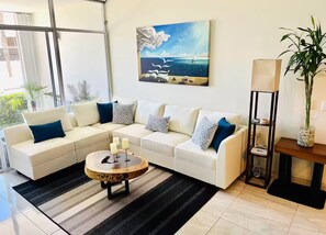 Een moderne woonkamer met een hoekbank, een ronde salontafel, een schilderij van een zeilboot, een hoge vloerlamp en een potplant.