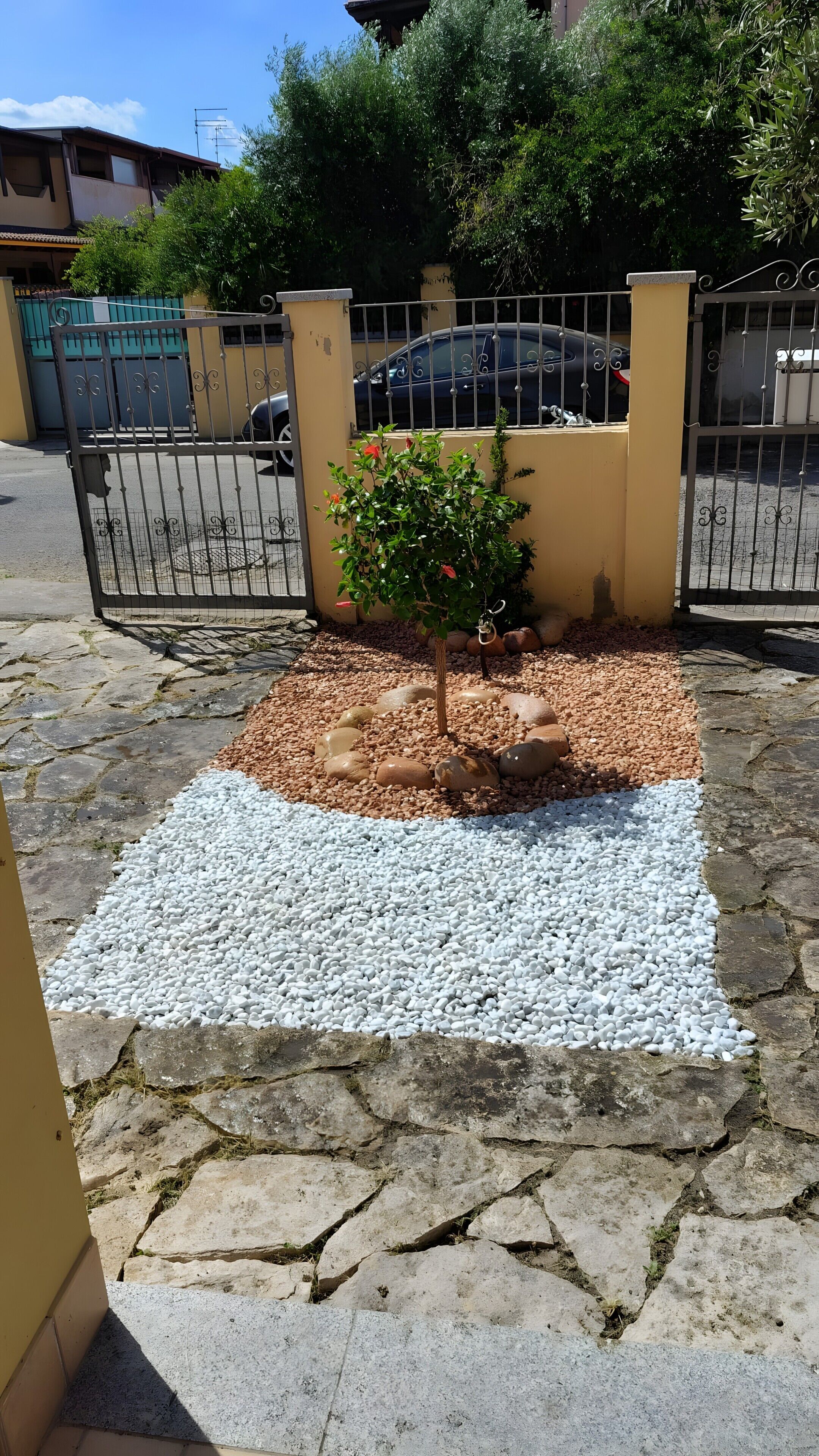 Uma área pavimentada com uma pequena árvore, pedrinhas e um portão de metal.