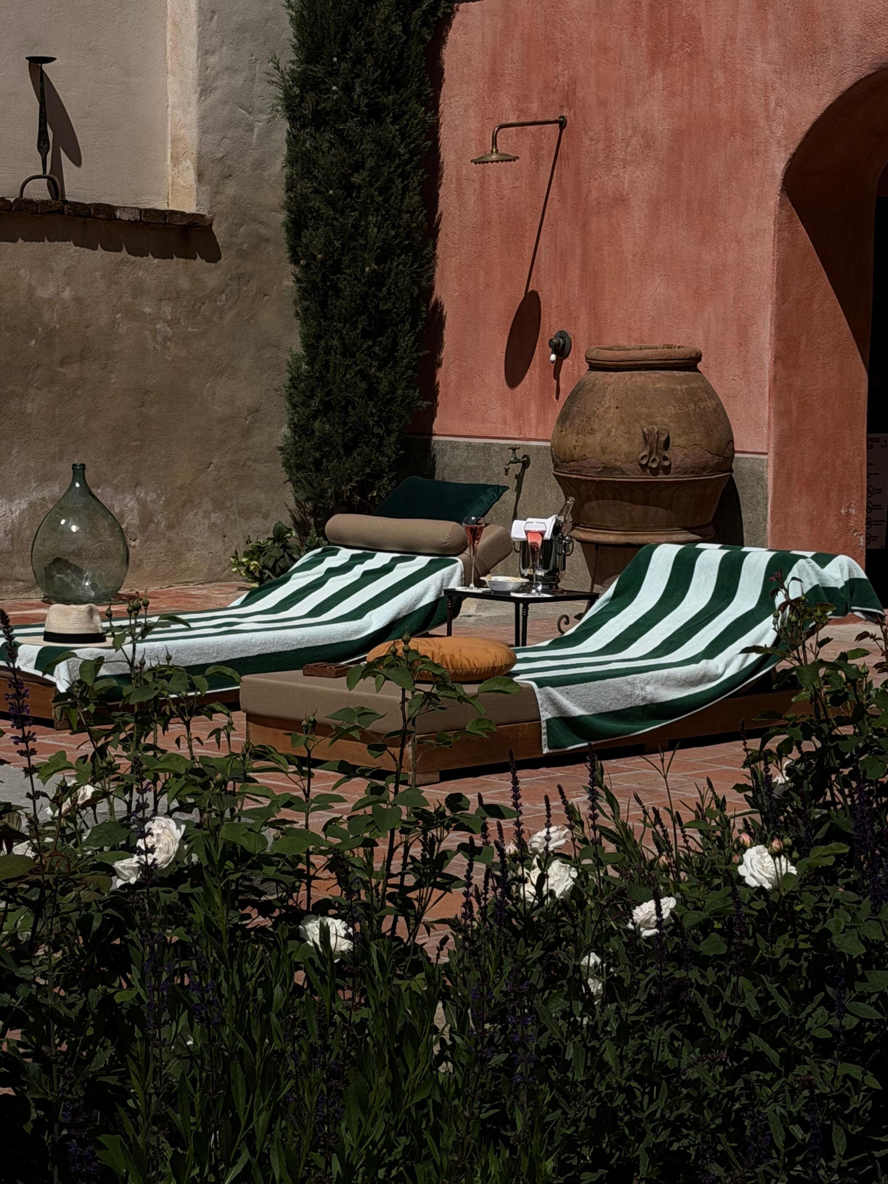 A sunlit outdoor lounge area with two striped lounge chairs, a large terracotta pot, and a wall-mounted showerhead.