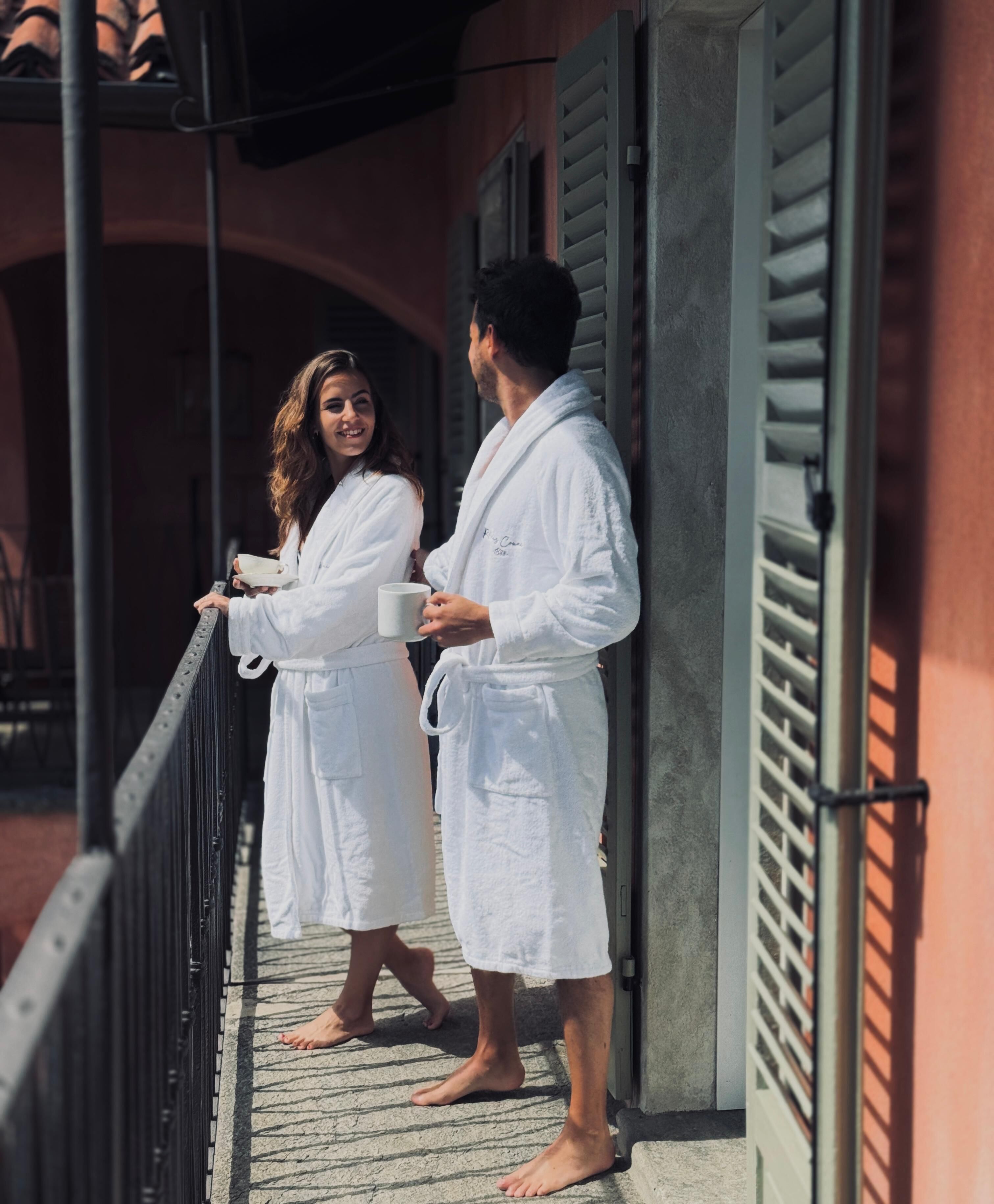 Two people in white bathrobes standing on a balcony with a railing, holding white mugs.