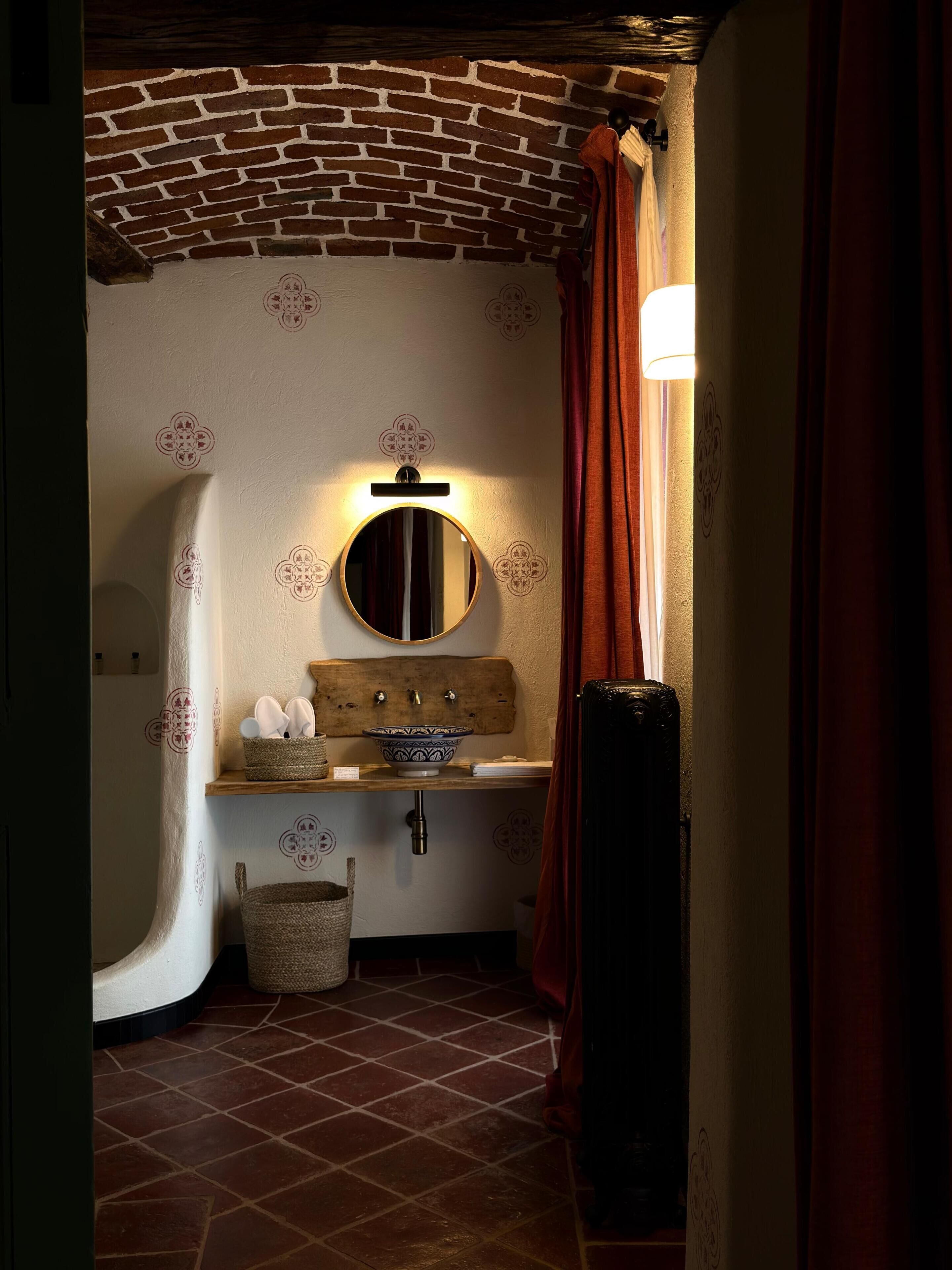Une salle de bain avec un plafond en briques, un miroir rond, un lavabo agrémenté d’un récipient décoratif et un porte-serviettes.