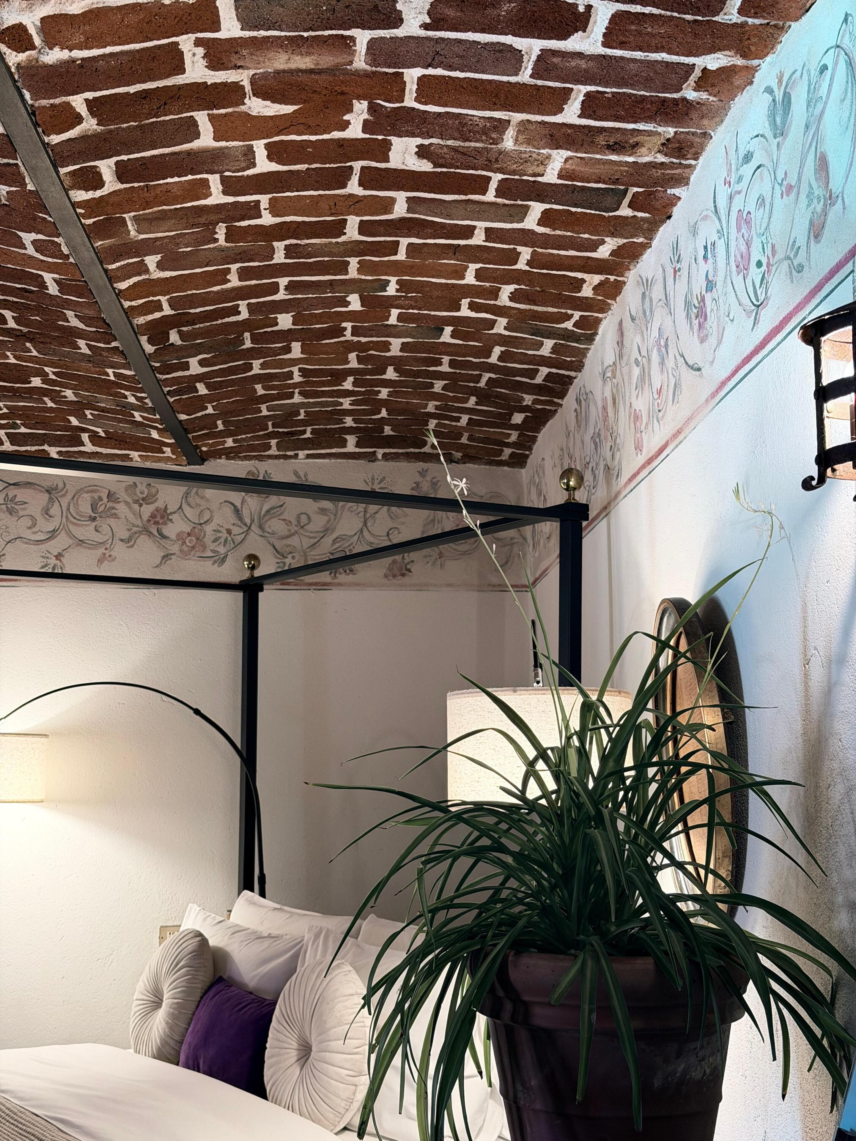 Een kamer met een bakstenen plafond, een bed met wit en paars beddengoed, een potplant en een muur met bloemenbehang.