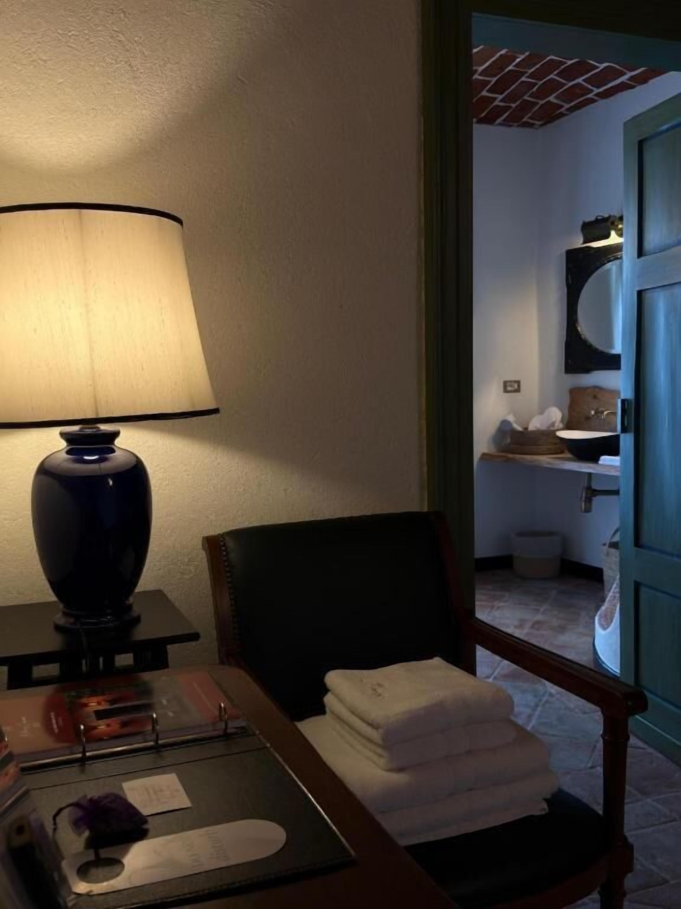 A hotel room with a desk, a lamp, a chair, a stack of towels, and a view of the bathroom.