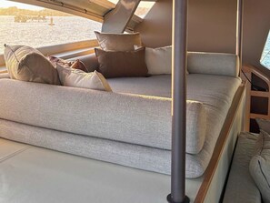 A cozy seating area with a cushioned bench and pillows, a window with a view of the water, and a metal pole. - Sun-Kissed Sailing Adventure in Córcega (Cuchery)