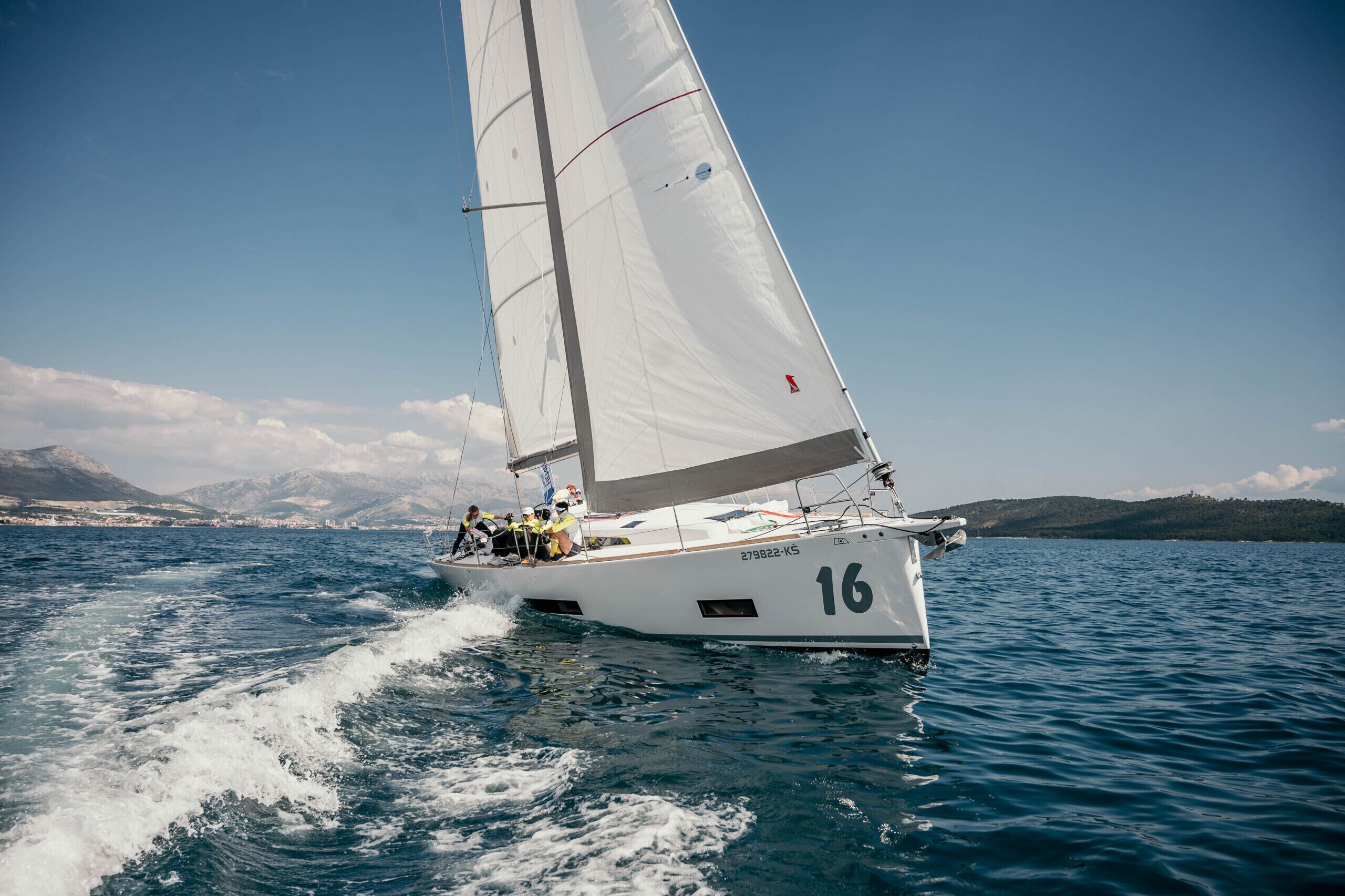 Eine Segelyacht mit der Nummer 16, die auf einem Gewässer mit einer bergigen Landschaft im Hintergrund fährt.