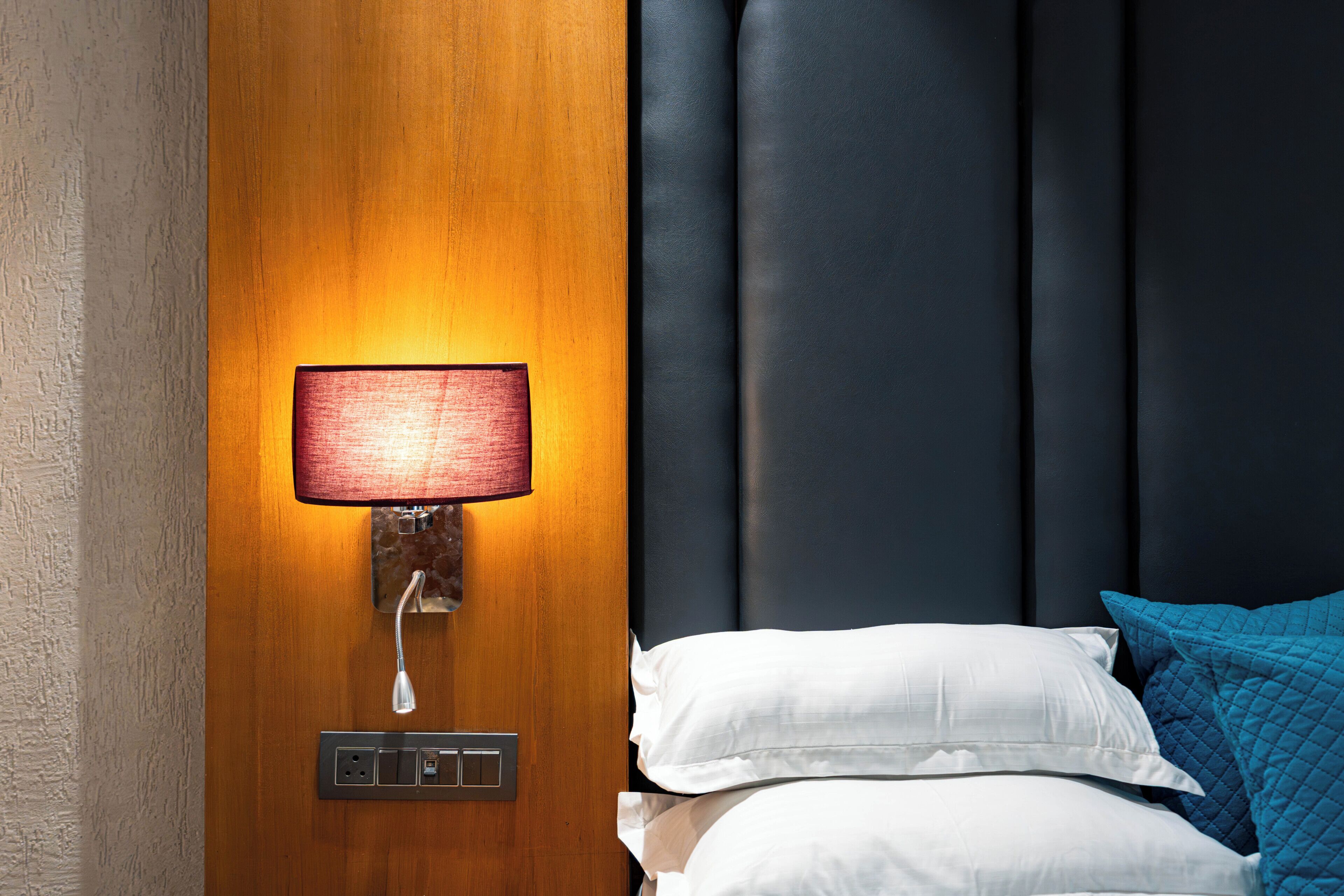 A hotel room with a wooden wall, a wall-mounted lamp, a bed with white and blue pillows, and a dark headboard.