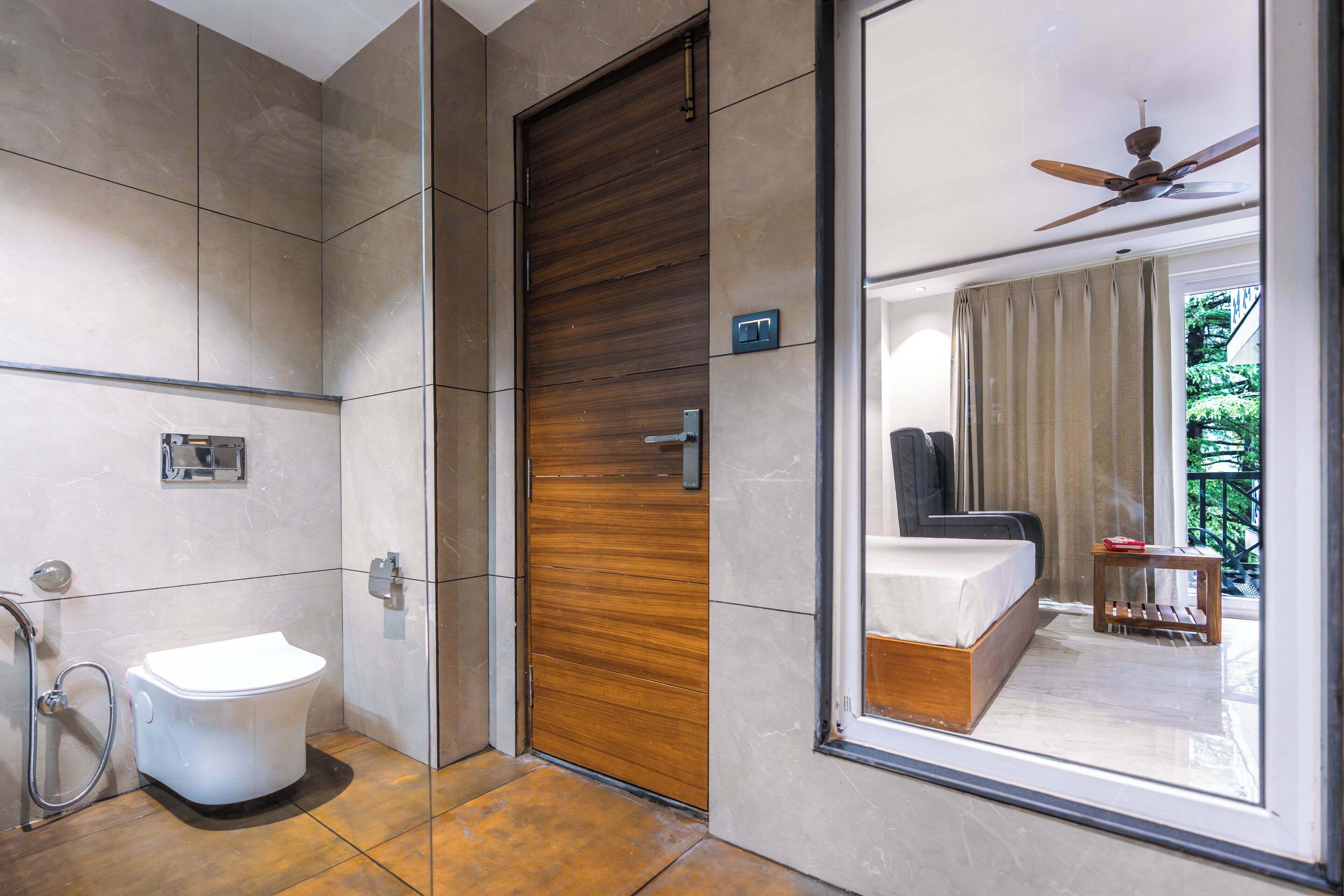 A modern bathroom with a toilet, shower, and a wooden door leading to a bedroom with a bed and a view of greenery.