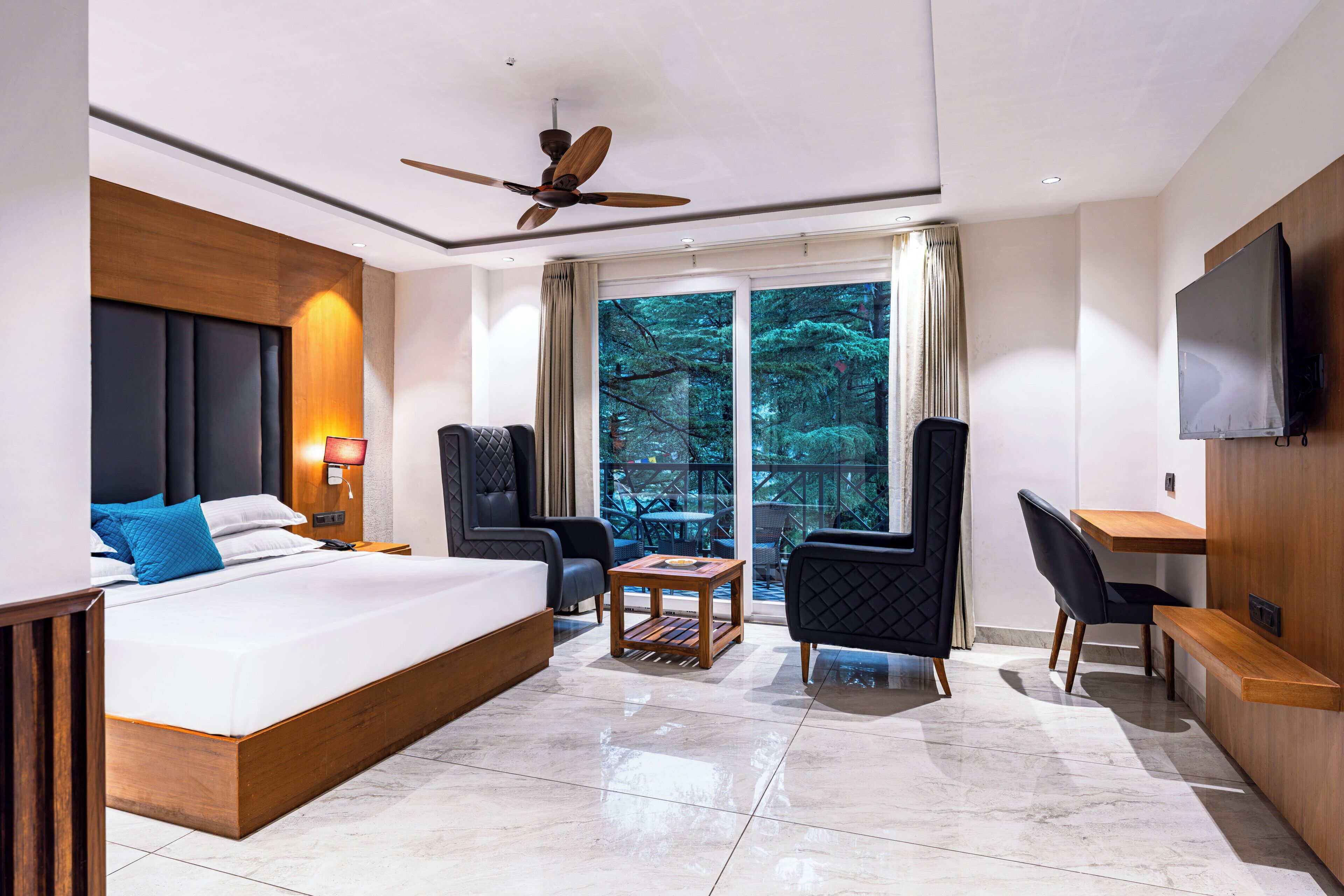 A modern hotel room with a large bed, a desk, two chairs, a ceiling fan, and a TV.