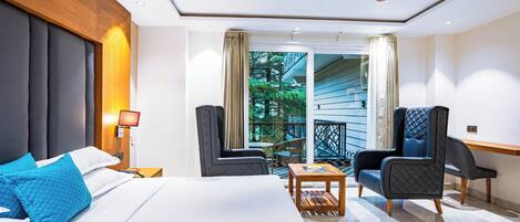 A modern hotel room with a bed, two armchairs, a small table, a ceiling fan, and a view of the outdoors.