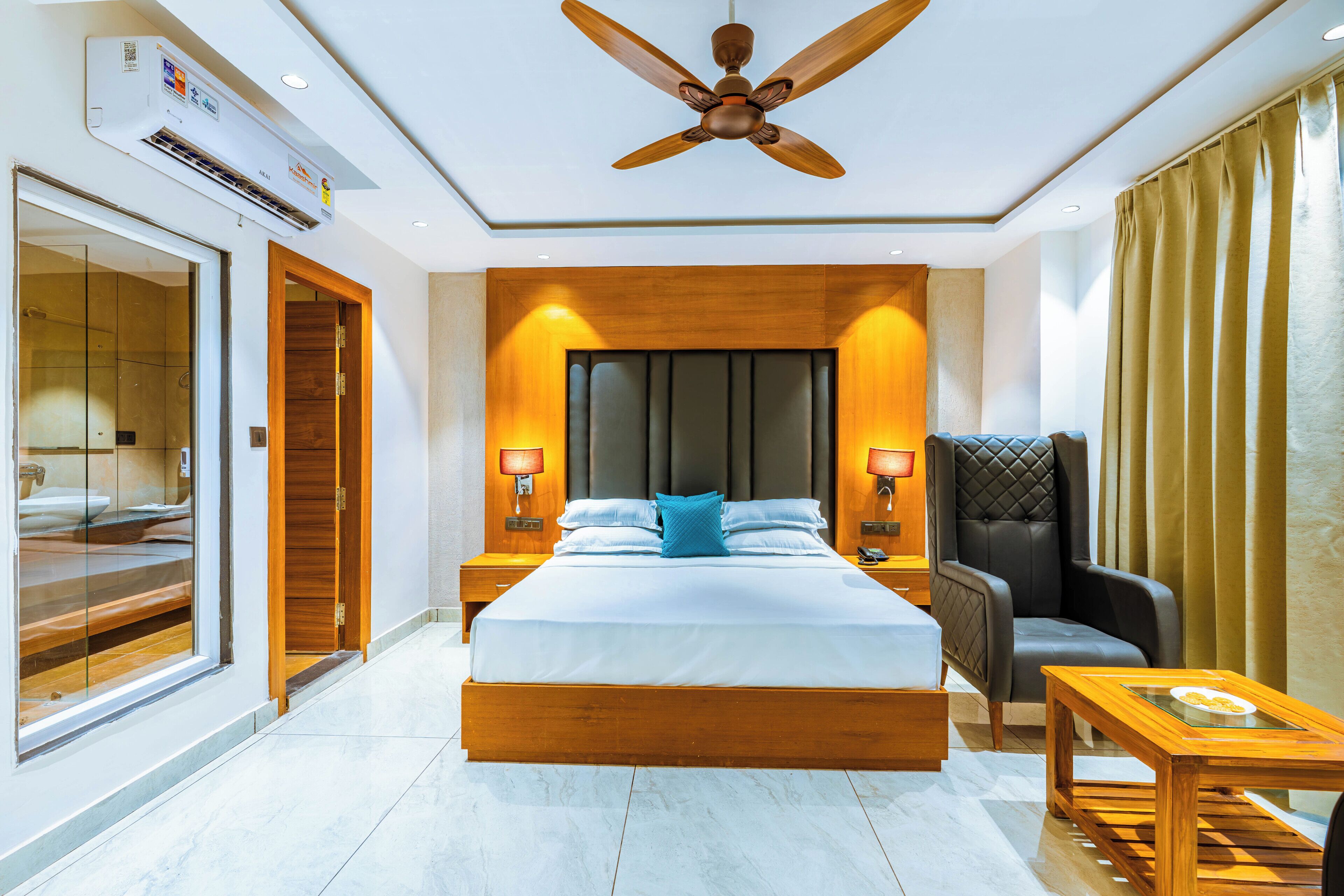 A modern hotel room with a large bed, a ceiling fan, a wooden nightstand, a leather armchair, and a small table.