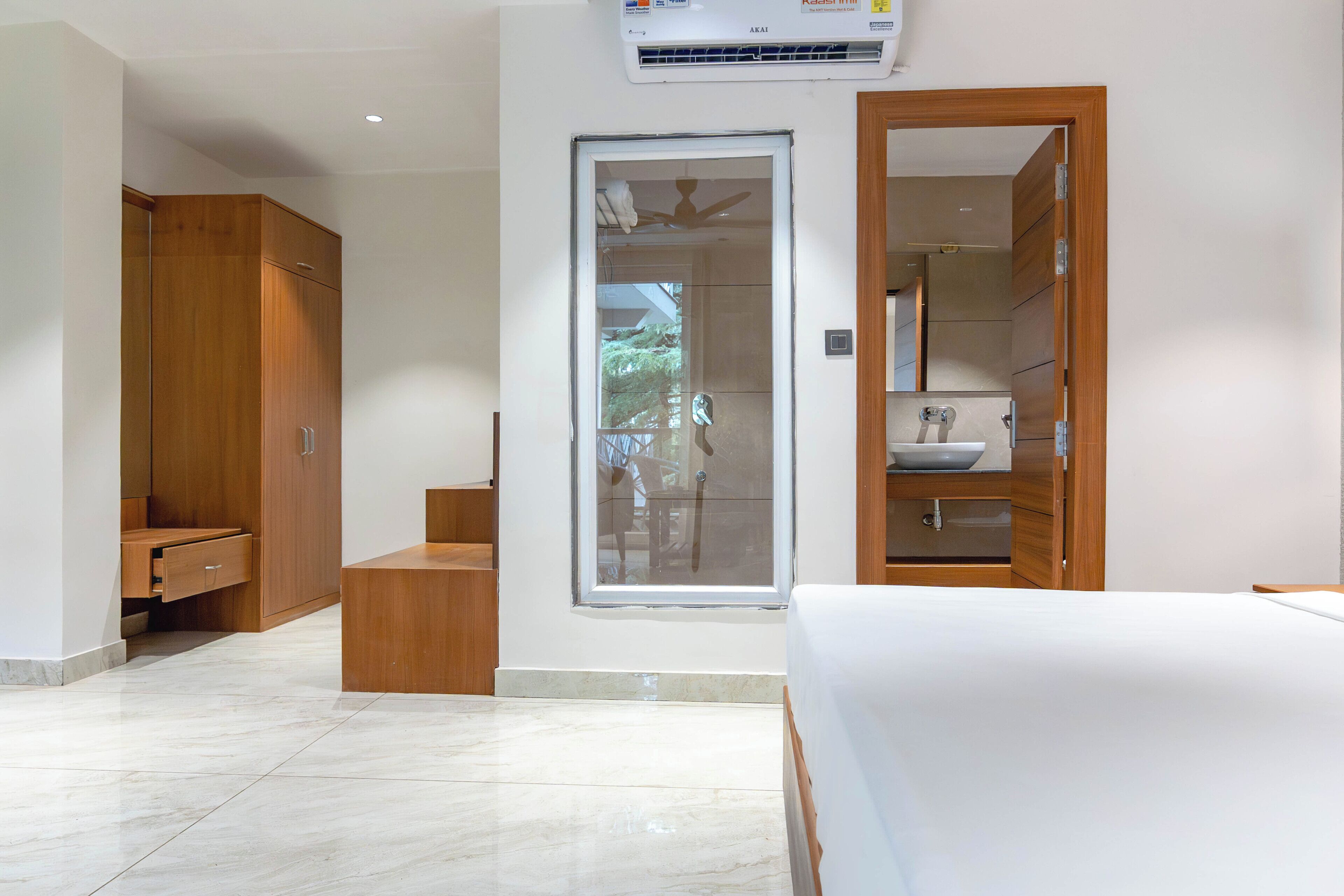 A modern bedroom with a large bed, a wooden wardrobe, a built-in bench, a glass door leading to a bathroom, and a ceiling fan.