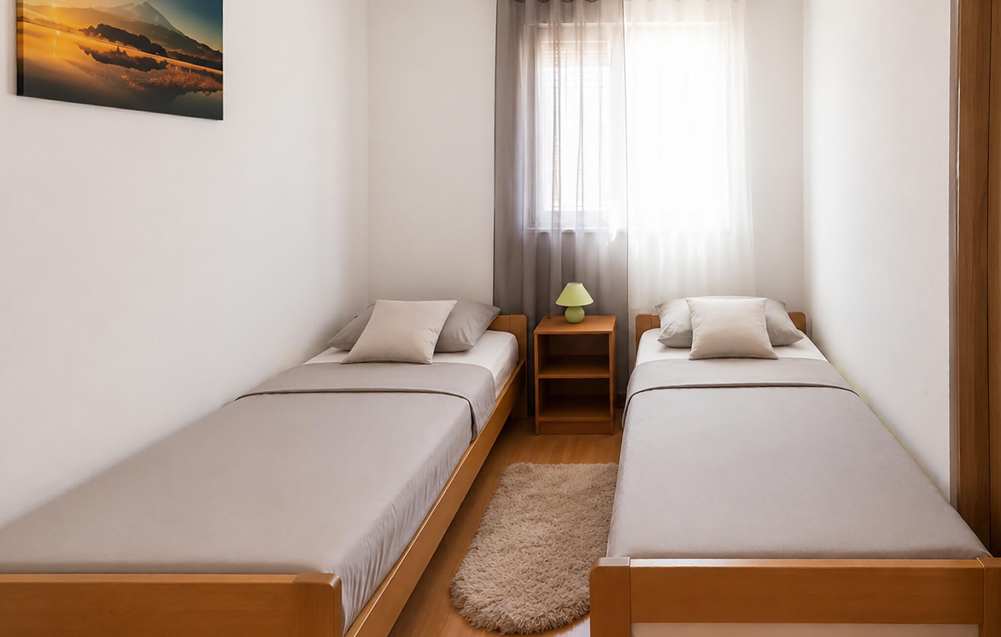 Una habitación pequeña y limpia con dos camas individuales, una mesita de noche con lámpara, una ventana con cortinas traslúcidas y un cuadro en la pared.