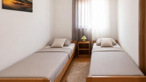 Ein kleiner, sauberer Raum mit zwei Einzelbetten, einem Nachttisch mit Lampe, einem Fenster mit durchsichtigen Vorhängen und einem Bild an der Wand.