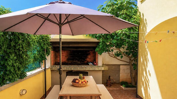 Une terrasse extérieure avec un four en briques, une table sur laquelle est posé un panier de fruits et un parasol.
