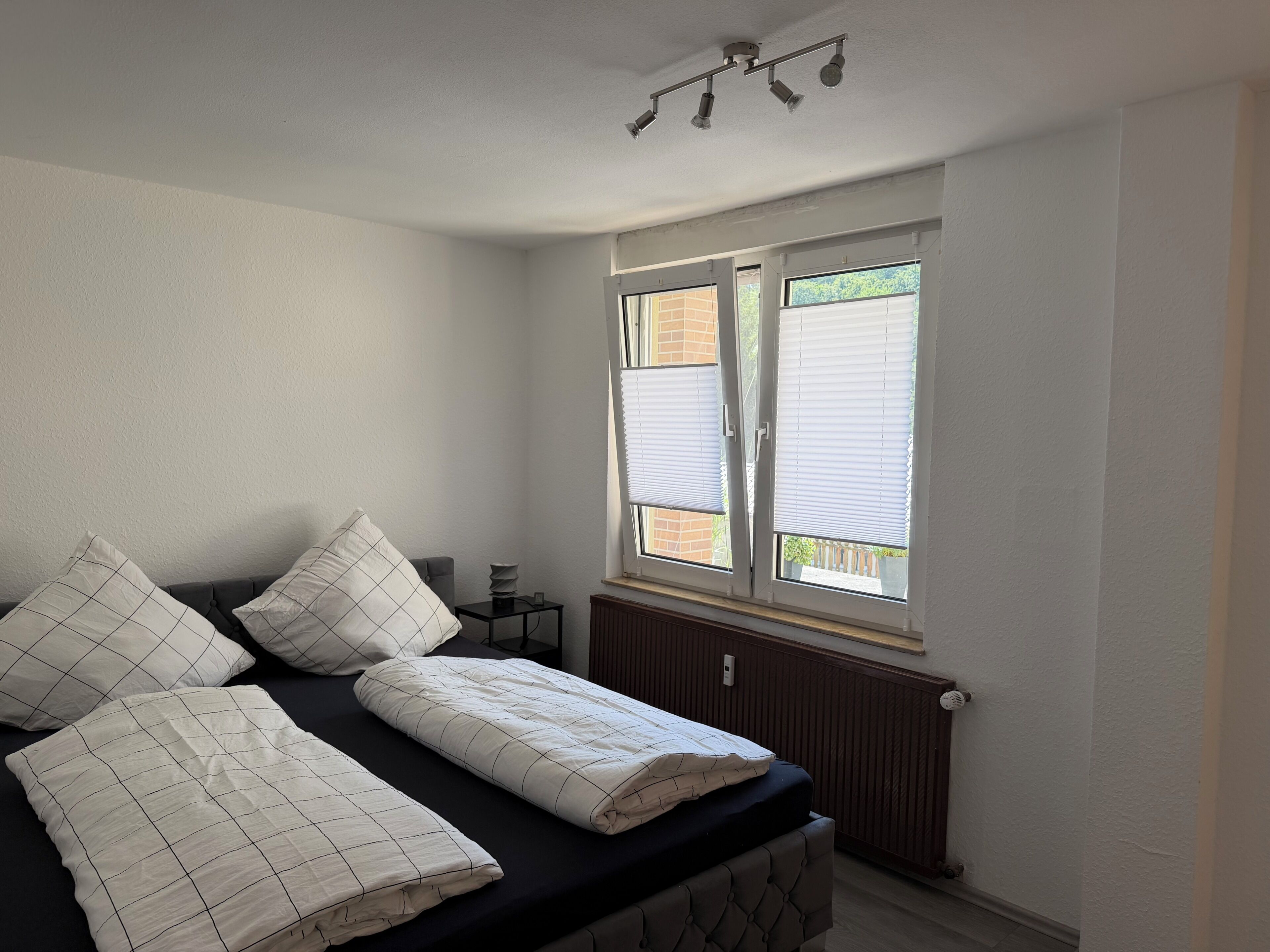 A bedroom with a bed, a window with blinds, a radiator, and a ceiling light fixture.