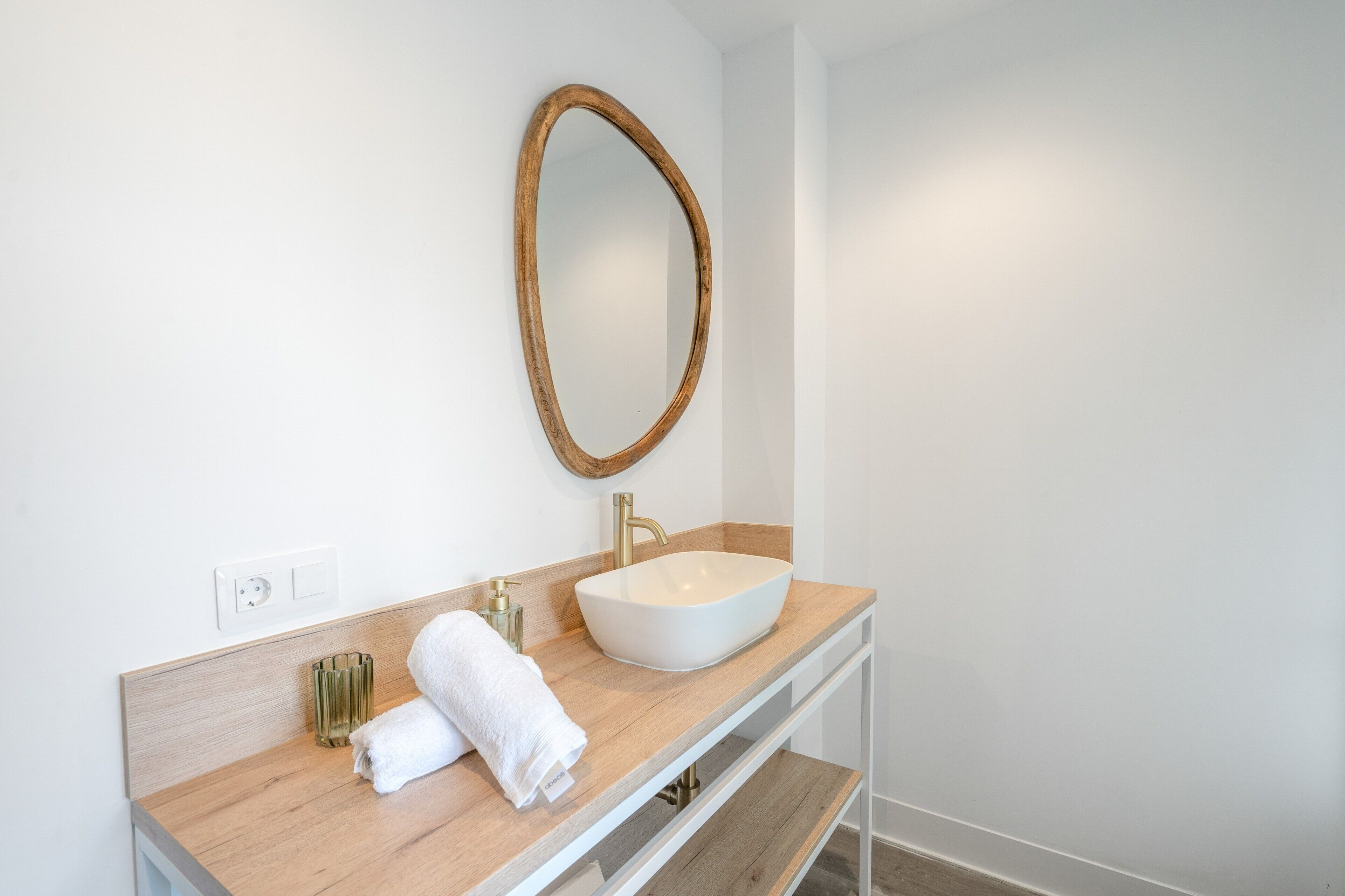 Un baño moderno con tocador de madera, un lavamanos blanco, un grifo dorado y un espejo con marco de madera.