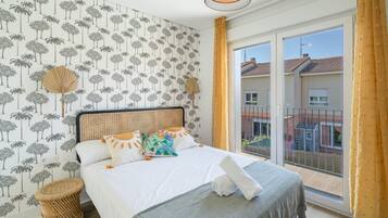 Een slaapkamer met een bed, nachtkastje, raam met gordijnen en uitzicht op gebouwen.
