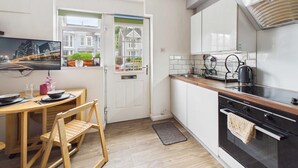 A compact kitchen with a dining area, a view through a door, and a modern stove. - The Mews - 1 Bed Holiday Home - Langland (Swansea)